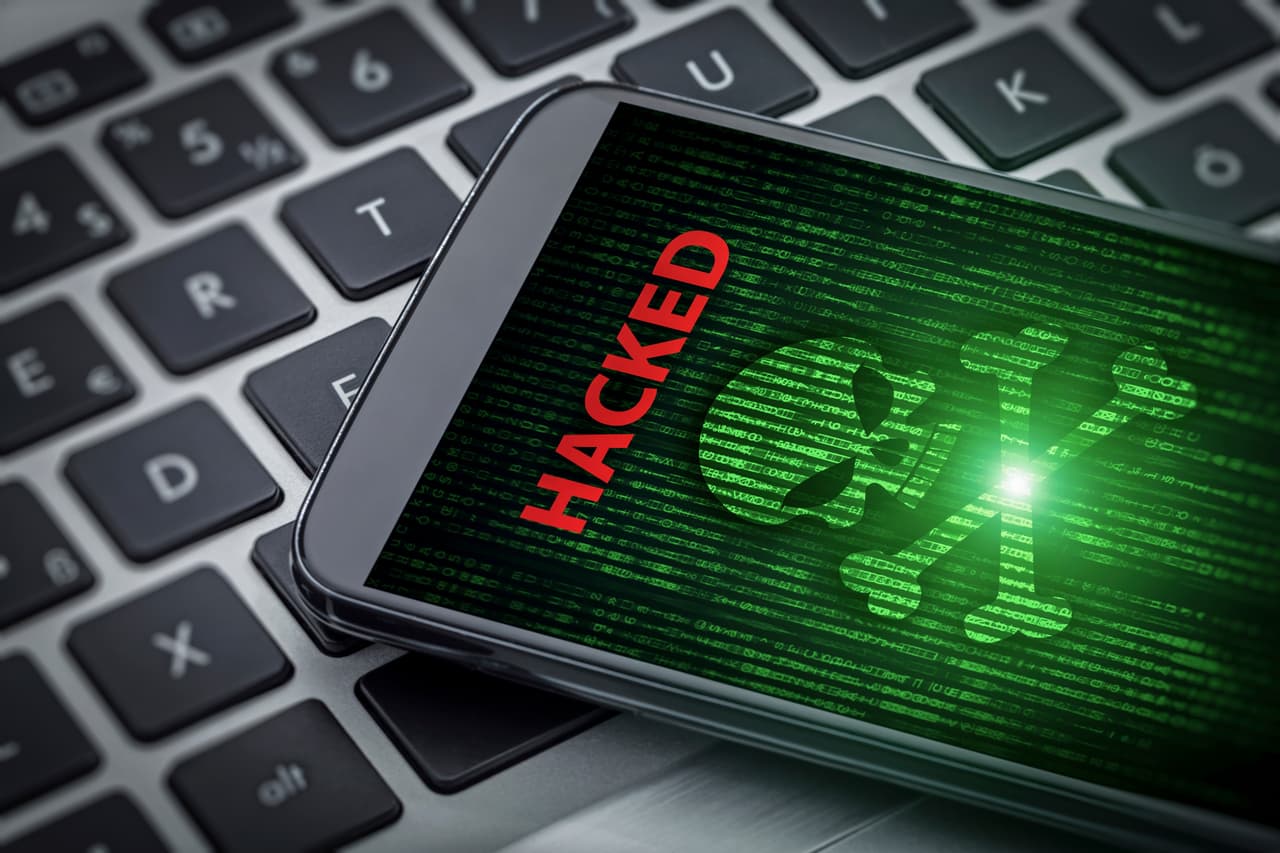 Hacked alert on the screen of smartphone on laptop computer. Skull of death on data flowing background. Selective focus on screen of the phone.